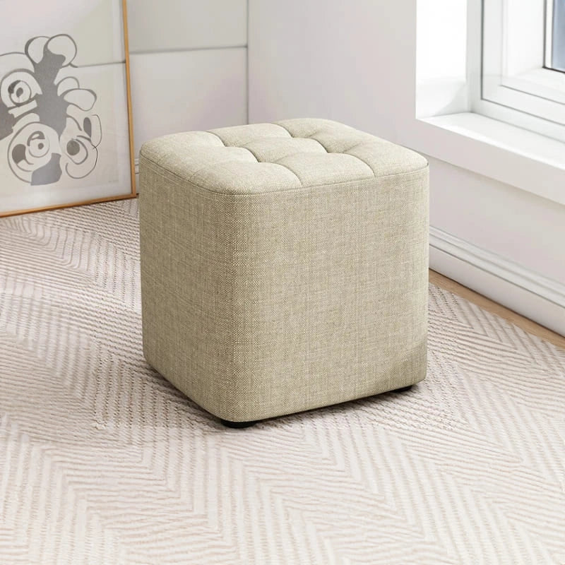 Beige cube ottoman on a patterned rug with a window and wall art in the background