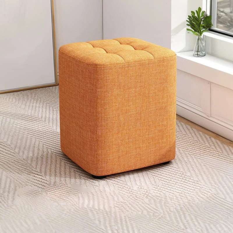 Orange cube-shaped ottoman on a carpeted floor with a neutral wall and plant in the background.