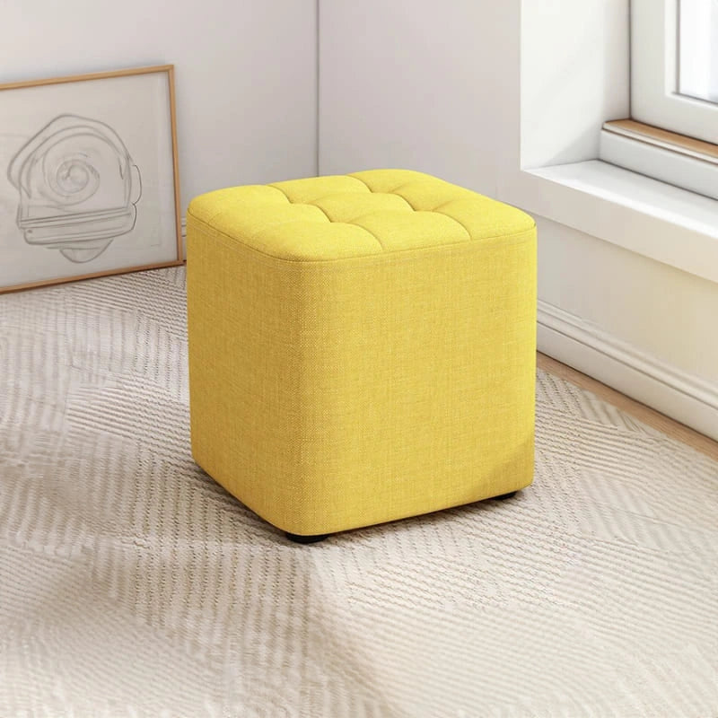 Yellow ottoman in a room with a window and framed picture on the wall.