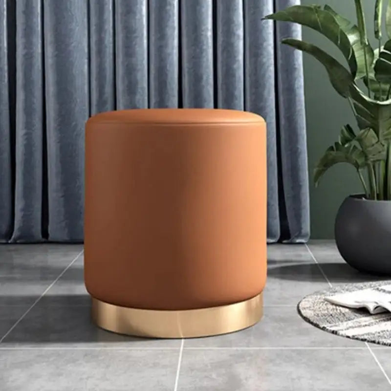 Brown ottoman with gold base on a gray tiled floor with curtains and plant in the background